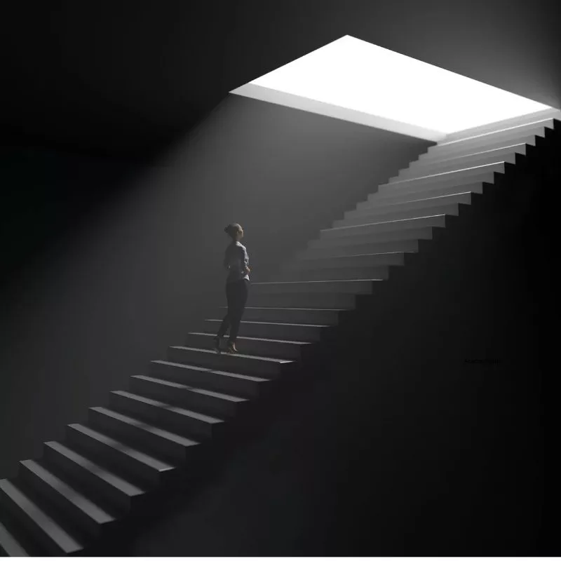 Person staring up a dark staircase to the light coming through above
